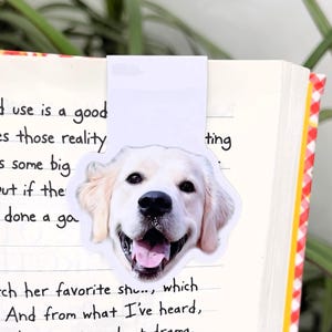 May include: A bookmark featuring a smiling golden retriever dog face. The bookmark is inserted into a book with handwritten text on the pages. The book has a red and white checkered pattern on the edge.