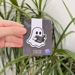 May include: A magnetic bookmark featuring a white ghost reading a book with the word "BOO" on it. The bookmark has a gray background with a bat and spiderweb design. The words "Magnetic Bookmark" are printed on the bottom.