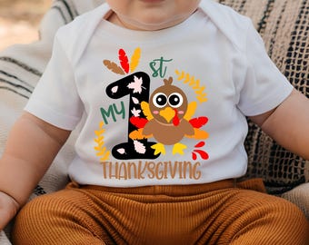 May include: A white short-sleeved baby bodysuit featuring a graphic of a brown turkey with colorful feathers, the text "MY 1st THANKSGIVING," and a large black number one decorated with small pink and white leaves, surrounded by yellow foliage.