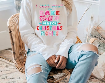 May include: A white long-sleeve sweatshirt featuring the colorful text I JUST WANT TO BAKE STUFF AND WATCH CHRISTMAS MOVIES in shades of pink, red, and teal. The garment has a crew neckline and ribbed cuffs, paired with light blue distressed jeans.