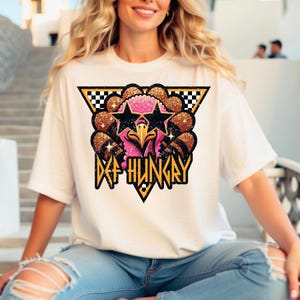 May include: A white short-sleeve t-shirt featuring a graphic of a turkey with pink skin and glittery brown feathers. The turkey wears black star-shaped sunglasses. The design includes a black and white checkered triangle and the text DEF HUNGRY.