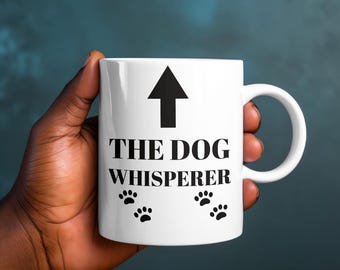 May include: White ceramic mug with a black arrow pointing upwards and the text "THE DOG WHISPERER" in black. Four black paw prints are below the text.