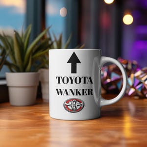 Può includere: Tazza in ceramica bianca con la scritta nera "TOYOTA WANKER", una freccia rivolta verso l'alto e il logo Toyota. La tazza è appoggiata su una superficie di legno, con piante sullo sfondo.
