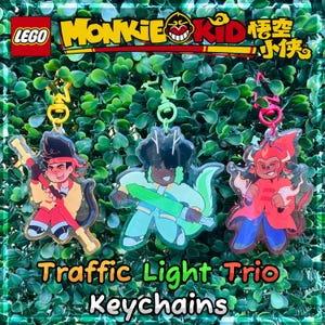 May include: Three acrylic keychains featuring characters from LEGO Monkie Kid. The keychains are in the shape of the characters, with vibrant colors including red, yellow, blue, and green. The text "Traffic Light Trio Keychains" is displayed at the bottom.