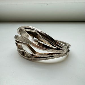 May include: A silver bracelet with a unique, abstract design. The bracelet is made of multiple interwoven bands that create a flowing, organic shape.