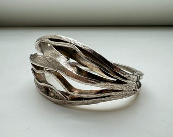 May include: A silver bracelet with a unique, abstract design. The bracelet is made of multiple interwoven bands that create a flowing, organic shape.