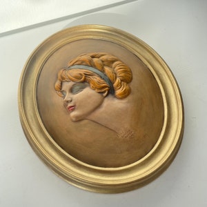 May include: A framed oval relief sculpture of a woman's head with a gold frame. The woman has red hair and is wearing a blue headband. The sculpture is signed "Ernst Guilbert Duizen 1910".