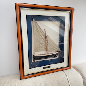 Op de afbeelding: Ingelijste nautische kunst met een gedetailleerd model zeilboot. De boot is zwart en wit met een beige zeil en houten mast. De lijst is een combinatie van zwart, goud en bruin, met een blauwe passe-partout. Een decoratief stuk voor een nautisch thema.