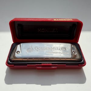 Könnte beinhalten: Ein silbernes Golden Melody-Mundharmonika in einem roten Etui. Auf dem Mundharmonika steht "Golden Melody" und "M. Hohner Made in Germany". Auf dem roten Etui steht das Wort "HANDMADE".