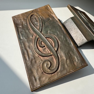 Puede incluir: Un porta-partituras de metal decorativo con una superficie texturizada y un diseño de clave de sol en relieve. El porta-partituras es de color bronce oscuro con una pátina verde.