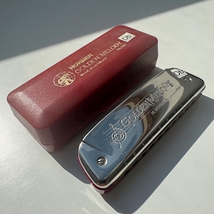 Pode incluir: Uma gaita Hohner Golden Melody prateada com estojo bordô. A gaita tem as palavras "Golden Melody" e um desenho de nota musical. O estojo tem o logotipo Hohner e as palavras "Golden Melody" e "Made in Germany".