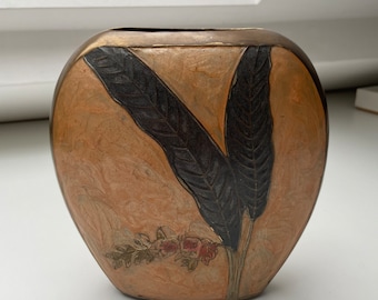 May include: A brass vase with a heart-shaped silhouette and a floral design. The vase has a marbled, gold-toned finish with black leaves and red flowers.
