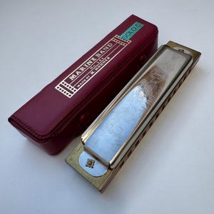 Könnte beinhalten: Eine silberne und goldene Mundharmonika mit einem weinroten Etui. Das Etui ist geöffnet und zeigt die Mundharmonika mit dem Text "MARINE BAND" und "MADE BY M. HOHNER". Die Mundharmonika hat aufwendige Gravuren.