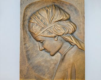 May include: A rectangular relief sculpture in sepia tones, showing a woman in profile with her head bowed. She wears a head covering, and her hair is pulled back. The artwork is set against a textured background, giving an antique appearance.