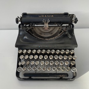 Puede incluir: Una máquina de escribir vintage negra con el nombre de la marca "URANIA" impreso en la parte superior. La máquina de escribir tiene un teclado QWERTY estándar con teclas etiquetadas con números y letras.