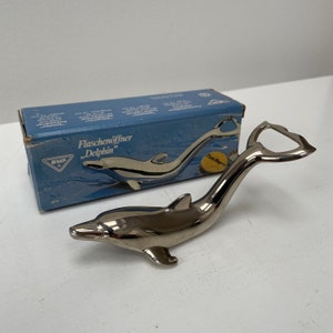 Può includere: Un apribottiglie a forma di delfino argentato con un'apertura a forma di cuore. L'apribottiglie è nella sua confezione originale, che è blu con testo bianco che dice "Flaschenöffner "Delphin"".