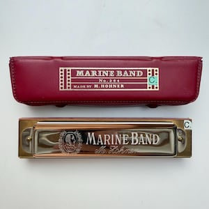 Könnte beinhalten: Eine silberfarbene Marine Band Mundharmonika von M. Hohner mit metallischem Finish und Gravur, begleitet von einem rechteckigen, dunkelroten Schutzetui mit einem goldbedruckten Etikett, das Marine Band No. 364 Made by M. Hohner liest.
