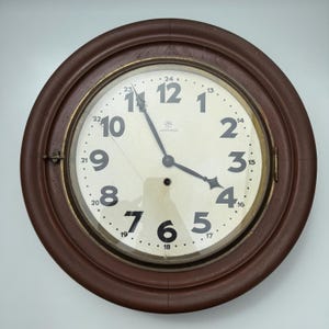 May include: A round, brown wooden wall clock with a cream-colored face. The clock features black numerals, black hands, and a glass cover. The clock's face has the word "Junghans" printed on it.