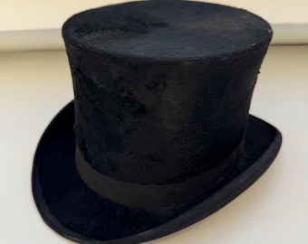 May include: A traditional black top hat with a cylindrical crown and a broad brim. It has a black ribbon band encircling the base of the crown. The hat is made of a soft, textured material.