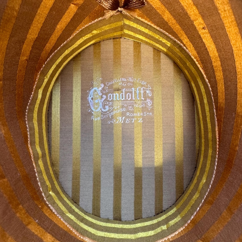 May include: Close-up of a vintage hat's interior, showcasing a striped pattern in gold and brown. The hat's lining includes a label with the name "Condolff" and the address "Rue Serpenoise 15, METZ".