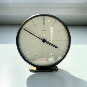 以下が含まれることがあります： クリーム色の文字盤と黒い針が付いた、ヴィンテージのKienzle時計。時計は濃いブロンズ色のフレームとベースを備えています。文字盤には時間を表す黒い線があります。クラシックな丸いデザイン。直径約15cm。