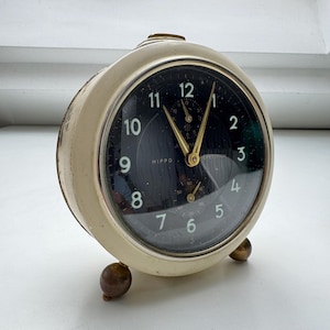 Puede incluir: Un reloj despertador redondo vintage con esfera negra y manecillas y números dorados. El reloj tiene una carcasa de metal color crema y descansa sobre dos pequeños pies redondos. La esfera del reloj dice "HIPPO".