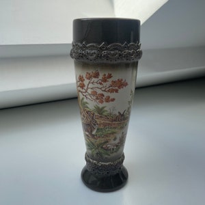 Puede incluir: Un jarrón decorativo y alto con la parte superior e inferior de color marrón oscuro y una banda de metal detallada. El jarrón presenta una escena pastoral con conejos, árboles y follaje en tonos marrón, verde y blanco.