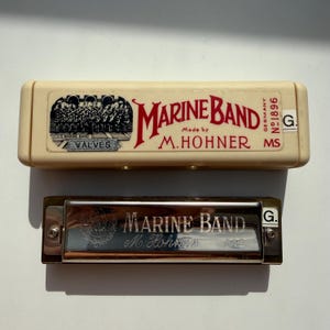 Könnte beinhalten: Eine Vintage Marine Band Mundharmonika und ihr Etui. Das Etui ist cremefarben mit rotem und schwarzem Text und einer Grafik einer Blaskapelle. Die Mundharmonika ist silberfarben mit den eingravierten Worten "Marine Band" und "M. Hohner".