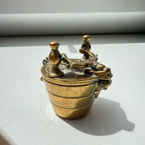 May include: A small, antique brass container with a lid. The container has a bucket-like shape with horizontal bands. The lid features two ornate handles and a decorative figure. The brass has a warm, aged patina.
