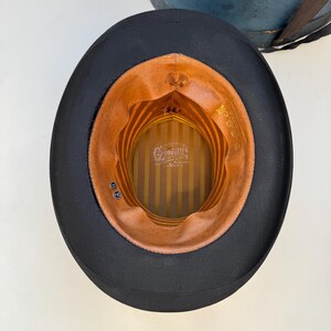 May include: A black top hat with a brown interior. The interior features a striped pattern and the brand name "Gandolfi's". The hat is likely made of felt or a similar material. The hat is a classic style.