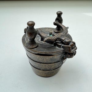 May include: A bronze-coloured, antique-style container with a lid. The container has a cylindrical body with horizontal bands. The lid features a dragon-like figure and three decorative knobs. The design suggests a vintage or historical artefact.