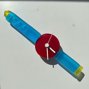 May include: A unique clock with a red circular face and white hands. The clock is attached to two blue glass rods with yellow tips.