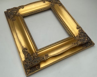 May include: A gold-colored ornate picture frame with a decorative, raised design. The frame is made of wood and has a rectangular shape.