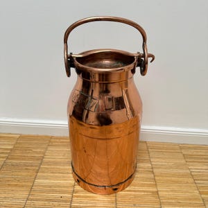 Puede incluir: Una antigua lechera de cobre con tapa abatible y asa de transporte. La lechera tiene el número "24507" estampado en el lateral.