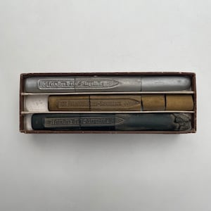 Puede incluir: Un juego de tres barras de cera para sellar vintage en una caja marrón. Las barras son plateadas, marrones y negras. La barra plateada tiene el texto "Feinster Brief-Siegelwachs" en ella. La barra marrón tiene el texto "Farblose Brief-Siegelwachs" en ella. La barra negra tiene el texto "Feinster Brief-Siegelwachs" en ella.