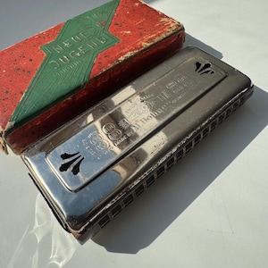 Pode incluir: Harmónica prateada vintage com o texto "M. Hohner" e "Neue Jugend" na caixa vermelha e verde. A harmónica tem recortes decorativos e é retangular. A caixa é vermelha com detalhes verdes.