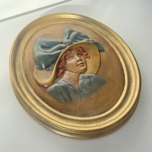 May include: A ceramic wall plaque with a gold frame depicting a woman with red hair wearing a blue hat with a large bow. The plaque has a textured, painted surface.