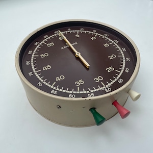 Op de afbeelding: Een witte en bruine analoge timer met een zwart gezicht en een witte naald. De timer heeft drie knoppen aan de zijkant, een rode, een groene en een witte.