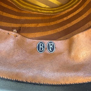 May include: Inside view of a hat featuring a brown leather sweatband and a striped brown and gold crown lining. Two circular metal badges with the letters "B" and "F" are visible on the sweatband.