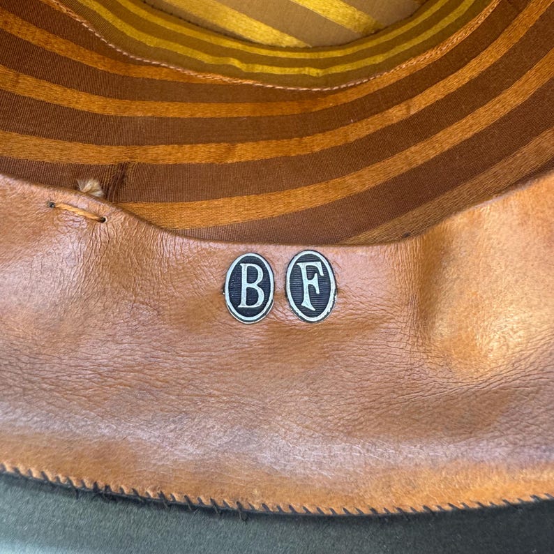 May include: Inside view of a hat featuring a brown leather sweatband and a striped brown and gold crown lining. Two circular metal badges with the letters "B" and "F" are visible on the sweatband.