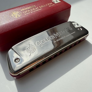 Könnte beinhalten: Eine silberne Hohner Golden Melody Mundharmonika in Fis-Dur mit rotem Kunststoffetui. Die Mundharmonika ist mit Golden Melody und M. Hohner Made in Germany sowie einem Violinschlüssel graviert. Das Etui ist mit F# No. 542 beschriftet.