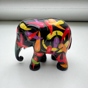 May include: A black elephant figurine decorated with colorful chili peppers. The peppers are red, yellow, orange, green, and purple. The elephant has a trunk, tusks, and four legs.