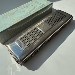 Könnte beinhalten: Eine silberne Vintage-Mundharmonika mit der Aufschrift "Bravi Alpini" und "M. Hohner Germany" liegt neben ihrer originalen hellblauen Schachtel. Die Mundharmonika hat eine rechteckige Form mit parallelen Schlitzen.