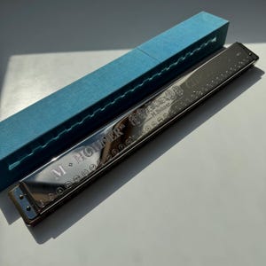 Könnte beinhalten: Ein silbernes M. Hohner Chromatic-Mundharmonika mit dem Text "CHROMATIC" und "M. Hohner" auf der Oberseite eingraviert. Das Mundharmonika befindet sich in einer blauen Box. Das Mundharmonika hat eine glänzende, reflektierende Oberfläche.