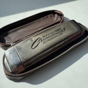 Puede incluir: Una armónica plateada M. Hohner Chromonica II De Luxe en un estuche marrón con cremallera. La armónica tiene los números del 2 al 12. El estuche está abierto, revelando un interior marrón.