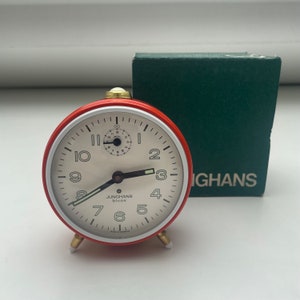 May include: A red and white alarm clock with a white face and black hands. The clock has a small second hand dial in the center. The clock is made by Junghans and says "bivox" on the face. The clock is sitting on a white surface.