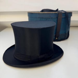 May include: A black top hat with a wide brim and a black band. The hat is on a white surface. A blue box with a leather strap is in the background.