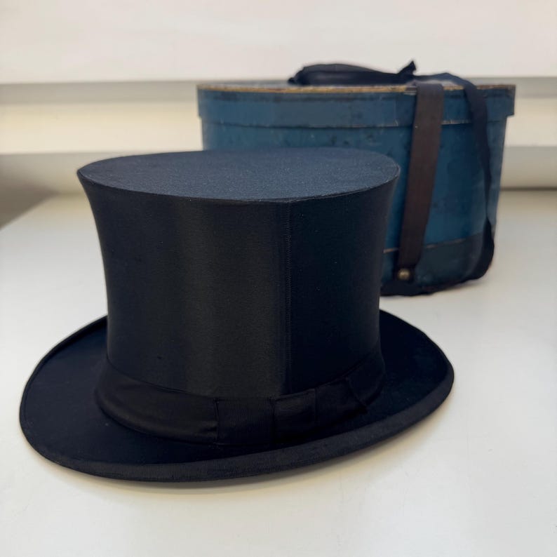 May include: A black top hat with a wide brim and a black band. The hat is on a white surface. A blue box with a leather strap is in the background.