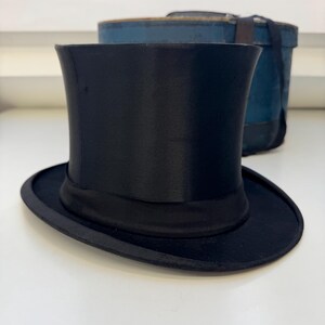 May include: A classic black top hat with a curved crown and a wide brim, featuring a black band. The hat is made of a shiny material. A blue box is in the background.
