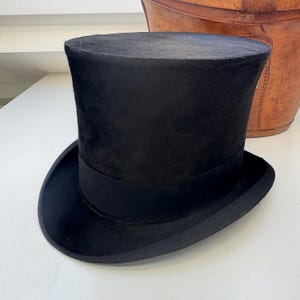 May include: A black top hat with a rounded crown and a wide brim. The hat has a black band around the base of the crown. A classic formal hat, often worn with formal attire.